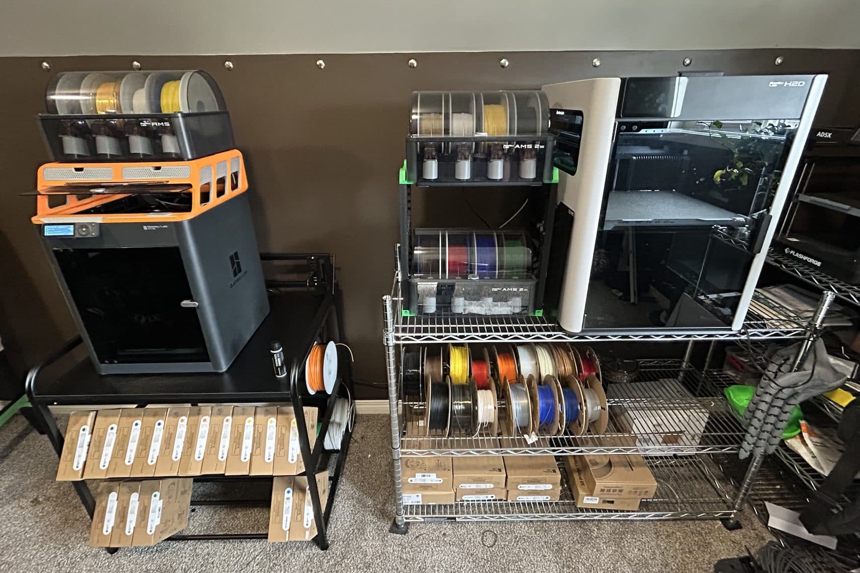 Bambu Lab H2D with two stacked AMS 2 Pro units, a second Bambu Lab printer with its AMS, and racks of filament spools and boxed rolls